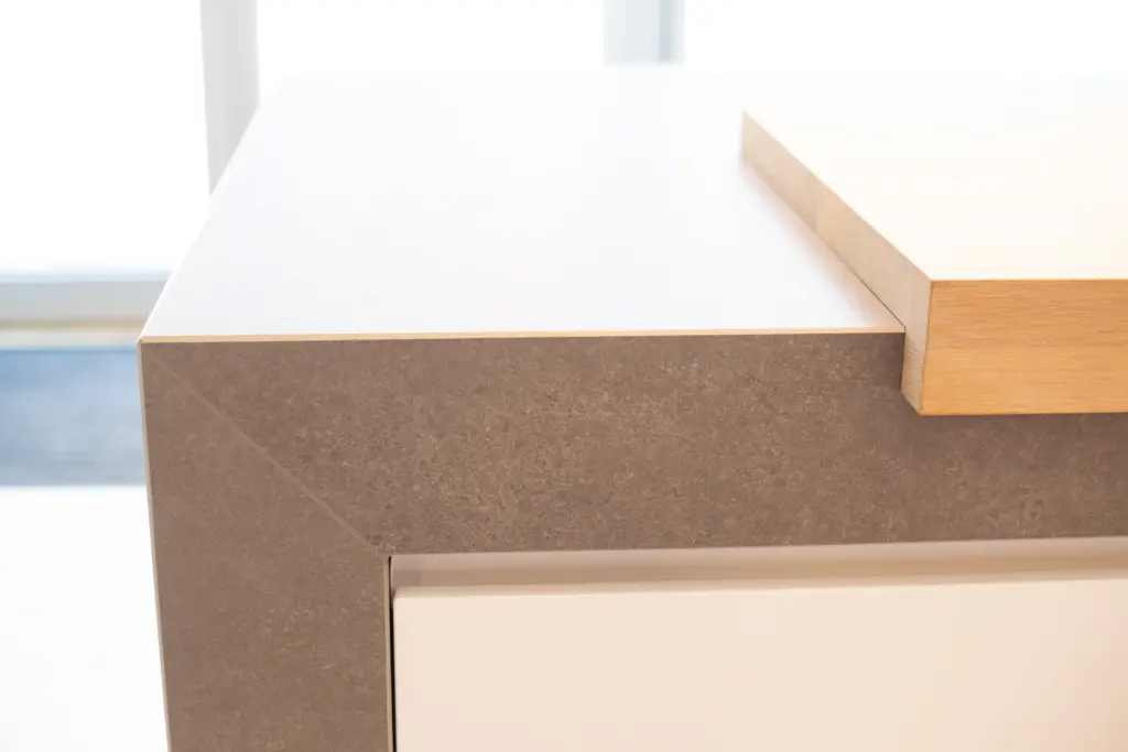Detail einer grauen Steinarbeitsplatte, deren Seitenwange auf Gehrung angesetzt ist, daneben ein Schneidbrett aus Holz