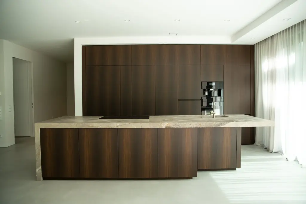 Kochinsel mit heller Steinarbeitsplatte über dunklen Holzfronten, dahinter eine Wand aus Hochschränken