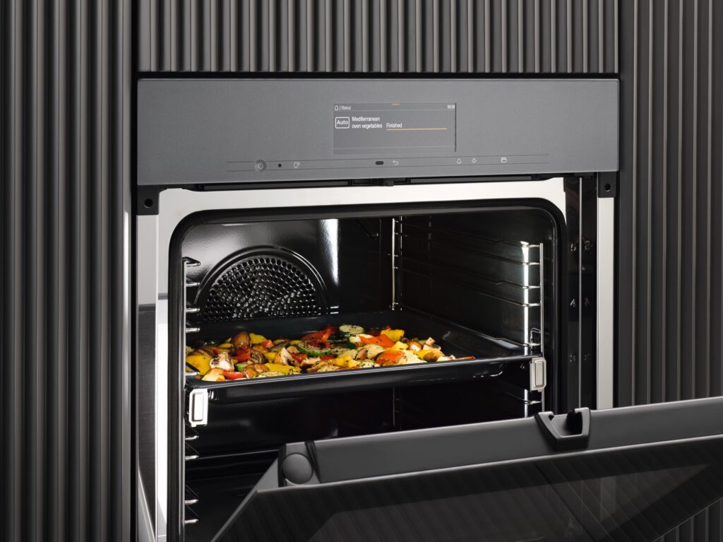 Geöffneter Miele Einbaubackofen in dunkler Front, auf der Tür ein Blech mit gebackenem Gemüse