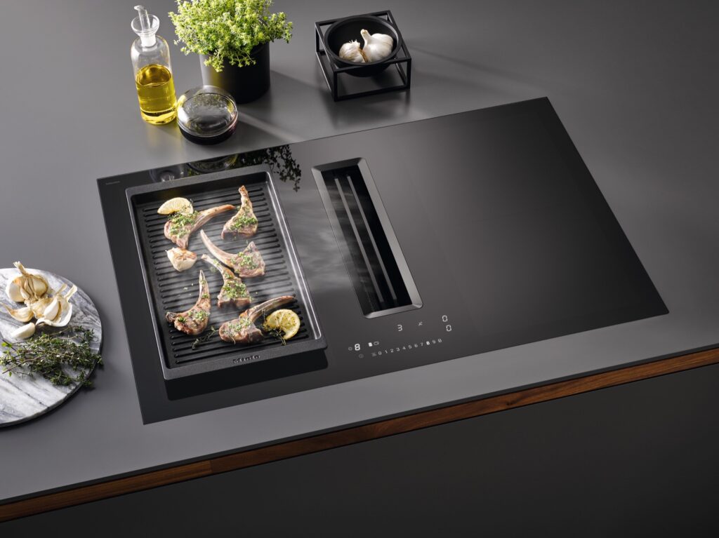Schwarzes Miele Induktionskochfeld mit mittig integriertem Dunstabzug und Grillplatte mit Gemüse