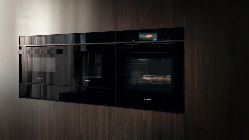 Zwei schwarze Siemens Einbaugeräte mit Glasfront nebeneinander in einer dunklen Holzwand, rechts ein beleuchteter Backofen